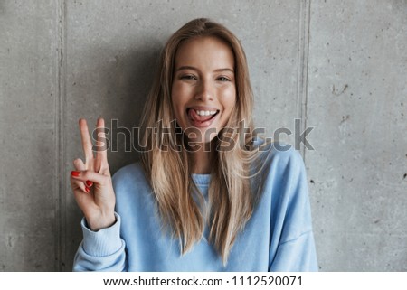 Picture of young cheerful happy pretty woman standing over grey wall background looking camera showing peace gesture.