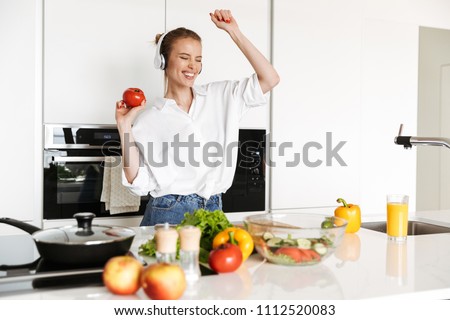 Picture of young amazing emotional woman dancing in kitchen indoors at home listening music with headphones cooking.