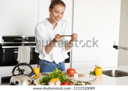 Picture of young beautiful woman standing in kitchen indoors at home using mobile phone take a photo cooking.