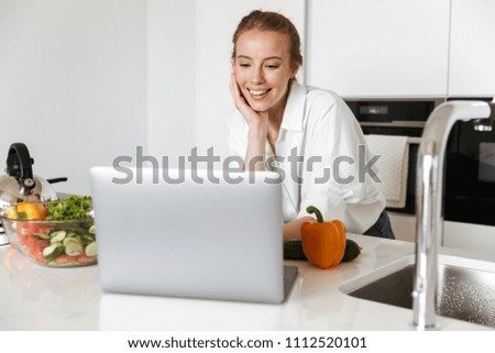 Picture of young pretty woman standing in kitchen indoors at home using laptop computer cooking.