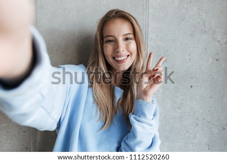 Picture of young cheerful happy pretty woman standing over grey wall background make selfie by camera showing peace gesture.