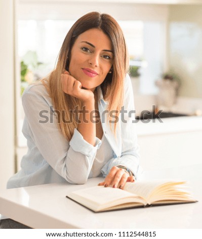 Beautiful young woman reading a book smiling and looking at the camera at home.