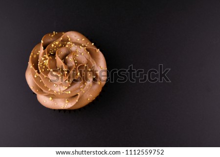Cupcake with whipped chocolate cream, decorated gold confectionery sprinkling on black background. Picture for a menu or a confectionery catalog. Top view.