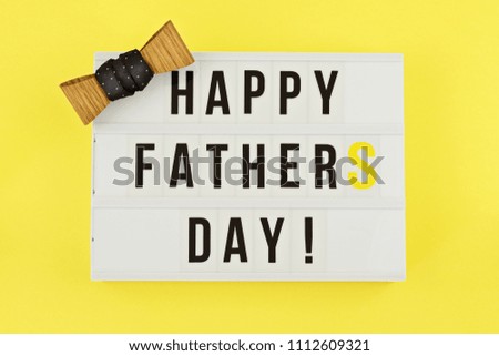 Happy Fathers Day text on white lightbox with trendy wooden tie bow on yellow background. Greeting card concept to celebrate Father's day
