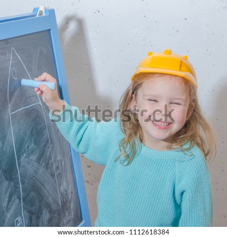 child engineer, designer, girl in a helmet paint a house on a black chalk Board