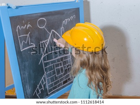 child engineer, designer, girl in a helmet paint a house on a black chalk Board