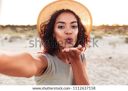 Close up of lovely young african girl in summer hat taking a selfie at the beach and sending kiss