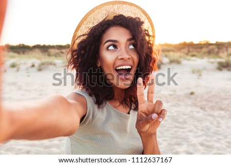 Close up of cheerful young african girl in summer hat taking a selfie at the beach and showing peace gesture