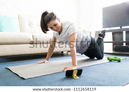 Portrait of attractive young latin woman learning new exercises watching online workout tutorials over her phone at home Royalty-Free Stock Photo #1112658119
