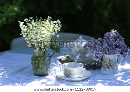 Floral scene with bouquet of the lily of the valley in vintage glass pitcher, lilac in china creamer, china pottery on linen napkin on table in garden, natural light and shadows, outdoor and space