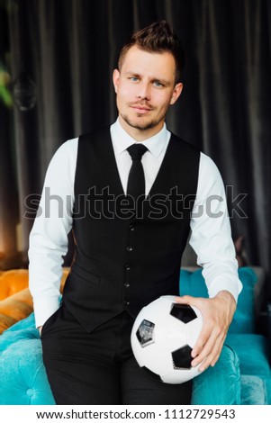 Stylish athletic man in a business costume vest holding a soccer ball. football player against the background of a interior cafe loft. Sits in the chair. Turquoise sofa. Hand in pocket of trousers.