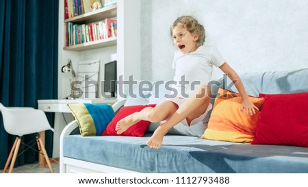 Little surprised and astonished teen girl watching tv at home. Female kid jumping on sofa