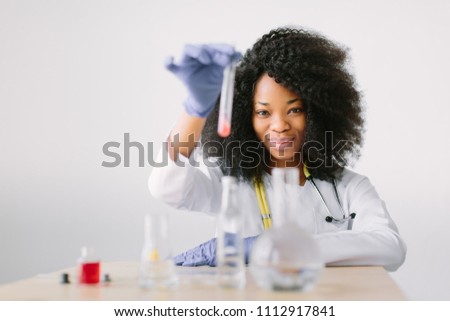Young beautiful African American girl doctor in a white coat with a stethoscope. sitting at a table with reagent flasks on white background. Investigator checking test tubes.