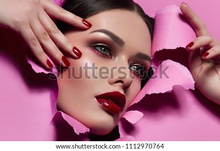 the face of a young beautiful girl with a bright make-up and with plump red lips peeks into a hole in pink paper.fashion, beauty, makeup, cosmetics, beauty salon, style, personal care, posture, hair.