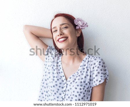 Redhead girl with pink rose in hair smiling in front of white wall.