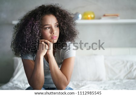 Lonely young latina woman sitting on bed. Depressed hispanic girl at home, looking away with sad expression. Royalty-Free Stock Photo #1112985545