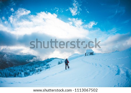 A man in snowshoes and trekking sticks in the mountains. Winter trip. Climbing climber against the background of sky with clouds. The tourist found shelter. House in the mountains. A hut in the snow.