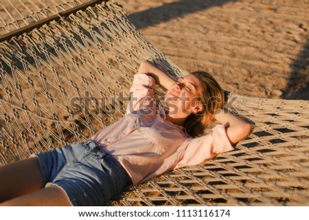 Young blonde girl wearing jeans shorts and lying on sand in wicker barefoot. Concept of summer vacations and resting on beach.