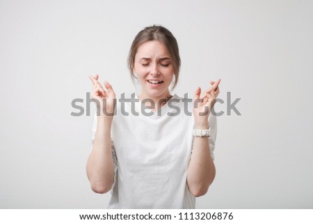 Indoor shot of attractive european girl in white shirt having excited look, keeping fingers crossed, eyes closed. She needs good luck before going to job interview. Positive facial emotion