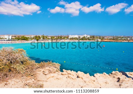 Image of a bay near Nissi beach, Agia Napa, Cyprus. Rough limestone coastline near deep blue transparent azure water, bush, and small houses on the background. Warm day in fall