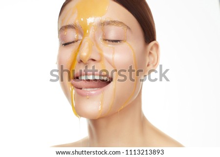 Close up beautiful women portrait, honey flowing down on her face and she is licking it. Clean perfect face. White background and studio light.