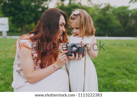 Joyful woman in light dress and little cute child baby girl take picture on retro vintage photo camera in park. Mother, little kid daughter. Mother's Day, love family, parenthood, childhood concept