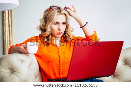 Young blonde woman work on red laptop while sitting on couch at home. Freelance and business for housewife