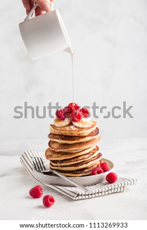 Stack of homemade pancakes with raspberries and pouring maple syrup on light marble background