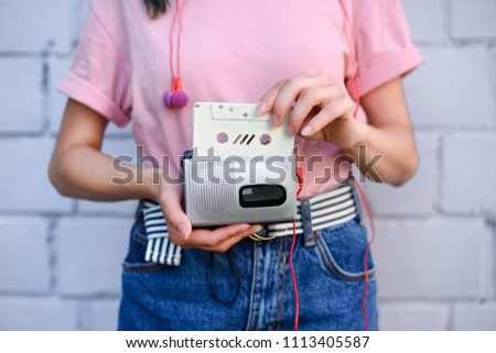 cropped shot of woman with earphones holding retro cassette player and audio cassette in hands against white brick wall
