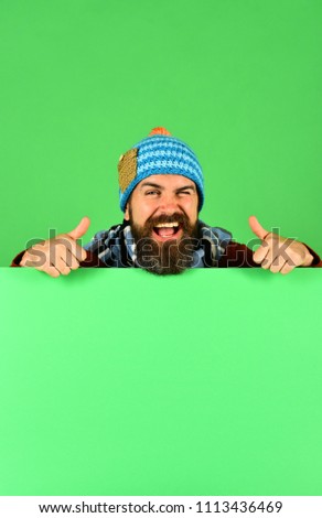 Autumn and demi season clothes concept. Man in warm hat shows thumbs up on green background, copy space. Hipster with beard and flirty face wears warm clothes. October and November sale idea