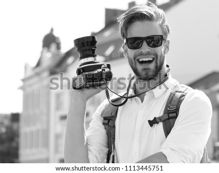 Urban photo and travelling concept. Tourist takes picture of cityscape. Young traveller or photographer with cheerful face goes sightseeing. Man with beard holds photocamera on urban background