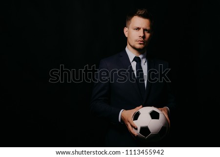 young handsome businessman holding a football on black background studio. blue jacket and tie. Stylish hairstyle model. World Championship. Royalty-Free Stock Photo #1113455942