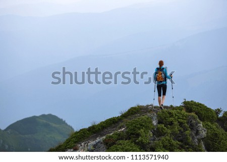 The tourist girl with the tracking sticks and the back sack at the edge of the cliff. The landscape with the high mountains. Endless vastness on the horizon. Warm sunny summer day.