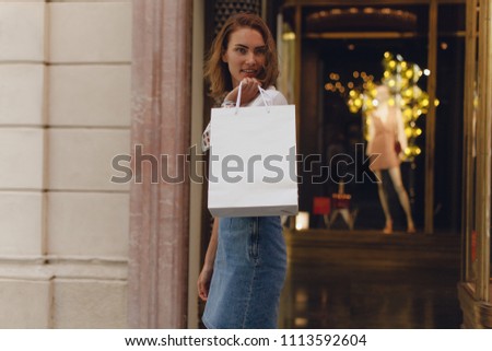 Beautiful happy young girl walking with shopping bag on city streets while doing shopping. Stylish girl smiling walking along clothes shop window. Mockup photo of white paper shopping bag