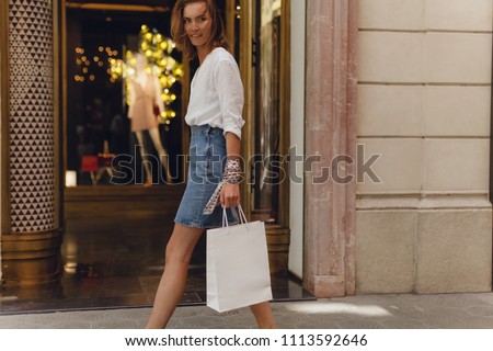 Attractive smiling girl walking with white blank shopping bag, doing shopping and buying clothes in shopping mall. Blurred clothes and dress on the background