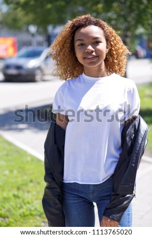 Young black woman with afro hairstyle smiling in urban park. Mixed girl wearing white t-shirt and blue jeans walking on the grass.