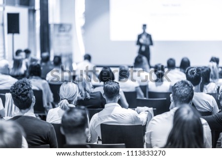Speaker giving a talk in conference hall at business event. Focus on unrecognizable people in audience. Business and Entrepreneurship concept. Blue toned greyscale image.