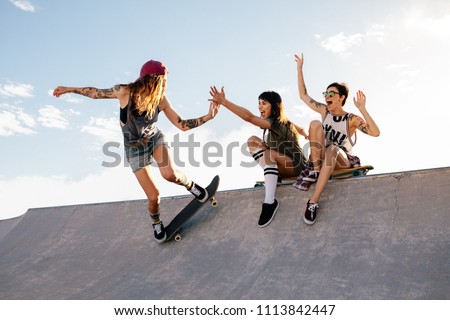 Female skateboarder riding skateboard at skate park with friends sitting on ramp having fun. Woman skater giving high five to female friend sitting on ramp during her routine.