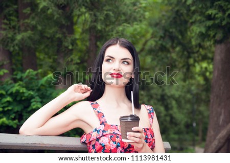 portrait of happy young pretty woman with dark brown hair drinks coffee on a bench on summer green park background 