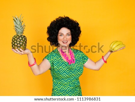 A girl in a bright green dress, pink pantyhose, pink beads and a pink bracelet. In a black wig of African style. Bright yellow background. She holds bananas and pineapple in her hands. Studio photo.