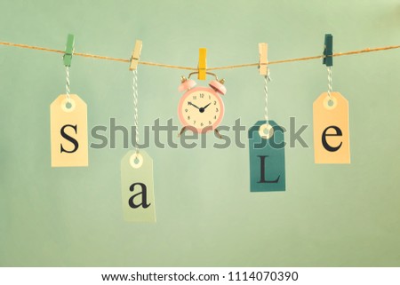 On a light blue background on the rope hang labels labeled SALE. In the center is a small pink alarm clock. Toned picture, concept, time for sales.