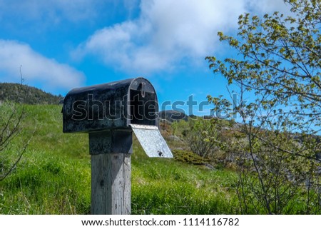 An antique country style black metal mailbox with the door open on a grey wooden post. The letterbox is under the blue sky and white clouds. There's a tree with leaves to the right of the mailbox. 