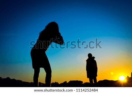Dark silhouettes of mother and daughter on the top of the hill on the clear sky background.  Mother takes pictures of daughter. Darkening vignette effect. Blurred background.