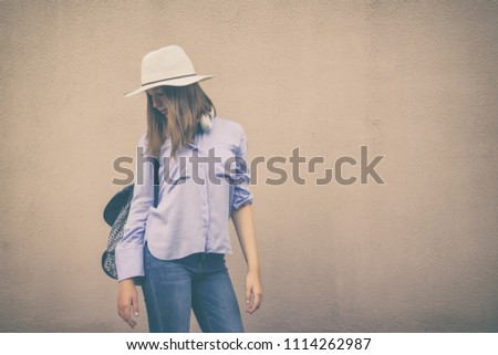 Young teenage tourist girl in straw hat, blue shirt and jeans stands with backpack and camera on wall background, portet.The concept of leisure and tourism in vintage style trend with copy space