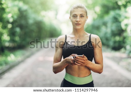 Woman Runner in the Summer Morning Park Listening to Music on Smartphone Using Bluetooth Earphones. Female Fitness Girl Jogging on Path Outside.