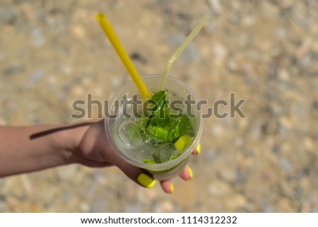 the maiden's hand holds a transparent glass of ice with lemon and mint. Mojito on the beach sand background