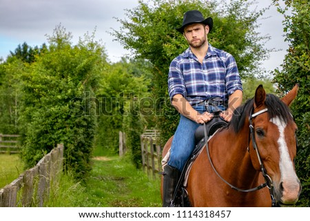 Good looking, hunky cowboy rides horse with boots, chequered shirt and hat Royalty-Free Stock Photo #1114318457