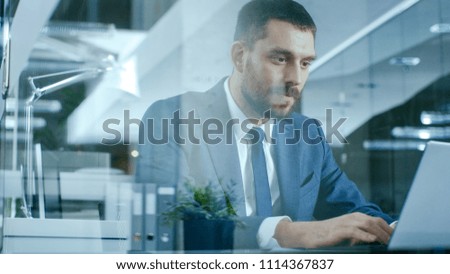Confident Handsome Businessman Works on a Laptop at His Desk. Stylish Man in Modern Glass Office.