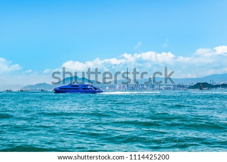 High speed water jet passenger nautical vessel moving fast from Hong Kong to Macau ( Asian gambling center). Bridge, cargo container port and terminal on background. Sunny day with blue cloudy sky. Royalty-Free Stock Photo #1114425200
