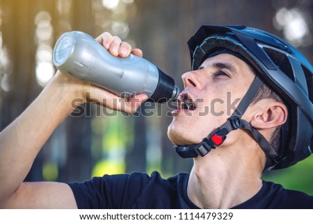 A cyclist in a helmet drinking water from a bottle while riding in the Park among the trees. Concept of active and healthy lifestyle